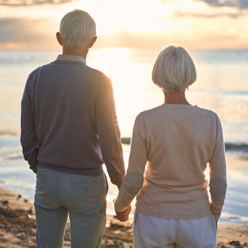 Elderly intelligent couple in love spending time romantically on the beach near the sea, walking holding hands and gently hugging.